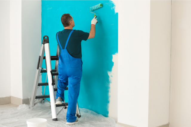 Worker dressed in blue overalls painting a wall blue on a ladder