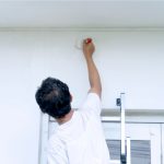 Worker painting exterior wall white on a ladder with a paint brush